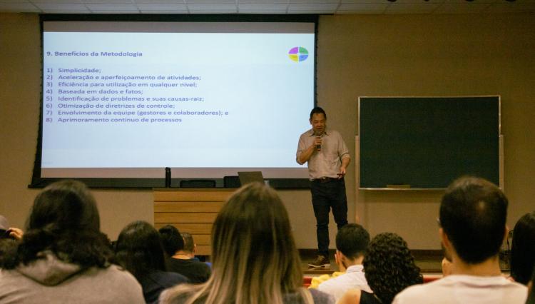 Palestrante de pé em frente a uma plateia em um auditório, apresentando um slide.