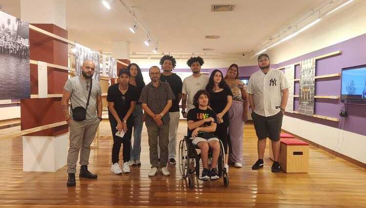 Grupo de estudantes e professores posando para uma foto no interior do Museu da Resistência, em São Paulo. Ao fundo, há painéis expositivos com imagens e informações sobre a Ditadura Militar no Brasil, enquanto o chão de madeira e a iluminação do ambiente destacam o espaço acolhedor e histórico do museu. 