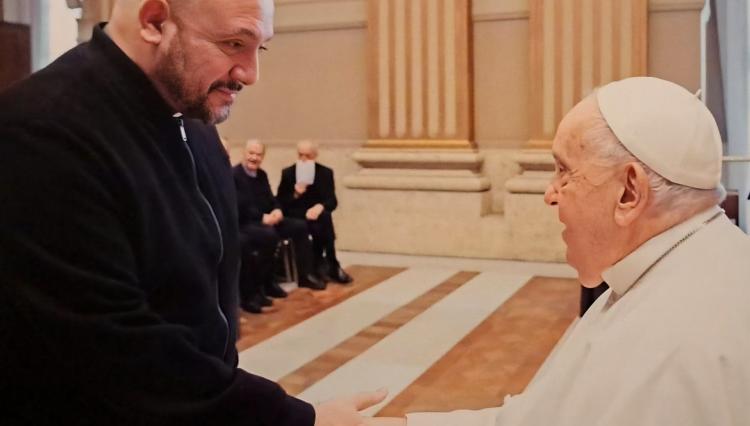 Fotografia de um momento significativo: o Professor Dr. Padre Rodrigo, de vestes escuras, aparentando ser um sacerdote, cumprimenta o Papa Francisco em um ambiente formal com arquitetura clássica. Ao fundo, outros clérigos estão sentados, observando a cena