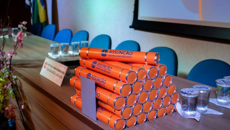 Canudos de diploma laranja com detalhes prateados, empilhados sobre uma mesa de formatura do Centro Universitário Assunção. Ao lado, copos de água mineral e um cartão de identificação. No fundo, cadeiras azuis, bandeiras e um telão.