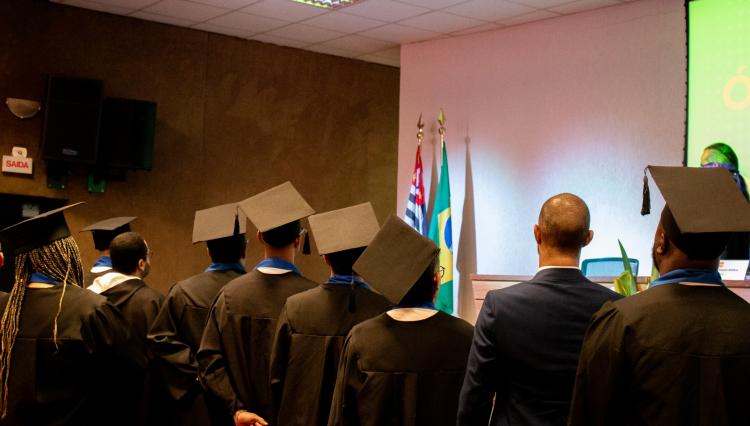 Formandos em becas pretas com capelos assistem a uma cerimônia de formatura em um auditório. Ao fundo, há bandeiras do Brasil e do estado de São Paulo, além de uma tela com parte de uma apresentação projetada. Um homem de terno está entre os formandos, virado para o palco