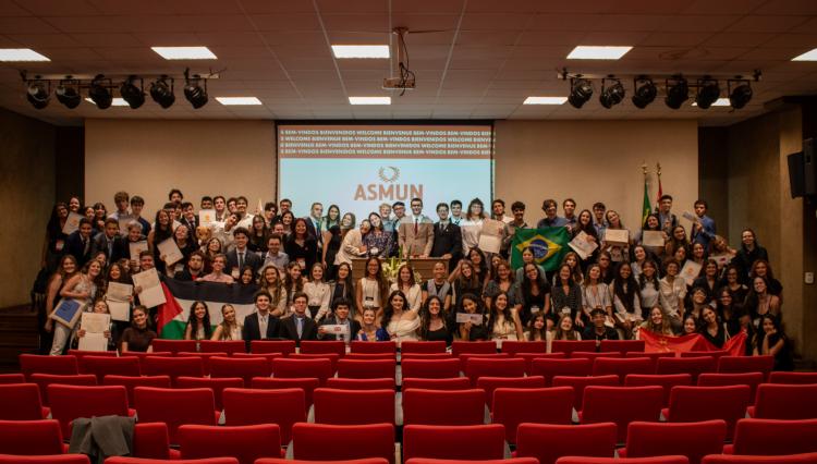  Um auditório repleto de estudantes participando do evento Assunção Model United Nations (ASMUN). Eles posam sorridentes para uma foto em grupo, segurando bandeiras, certificados e placas representando países e organizações. Ao fundo, um telão exibe o logo do ASMUN e mensagens de boas-vindas em diferentes idiomas.